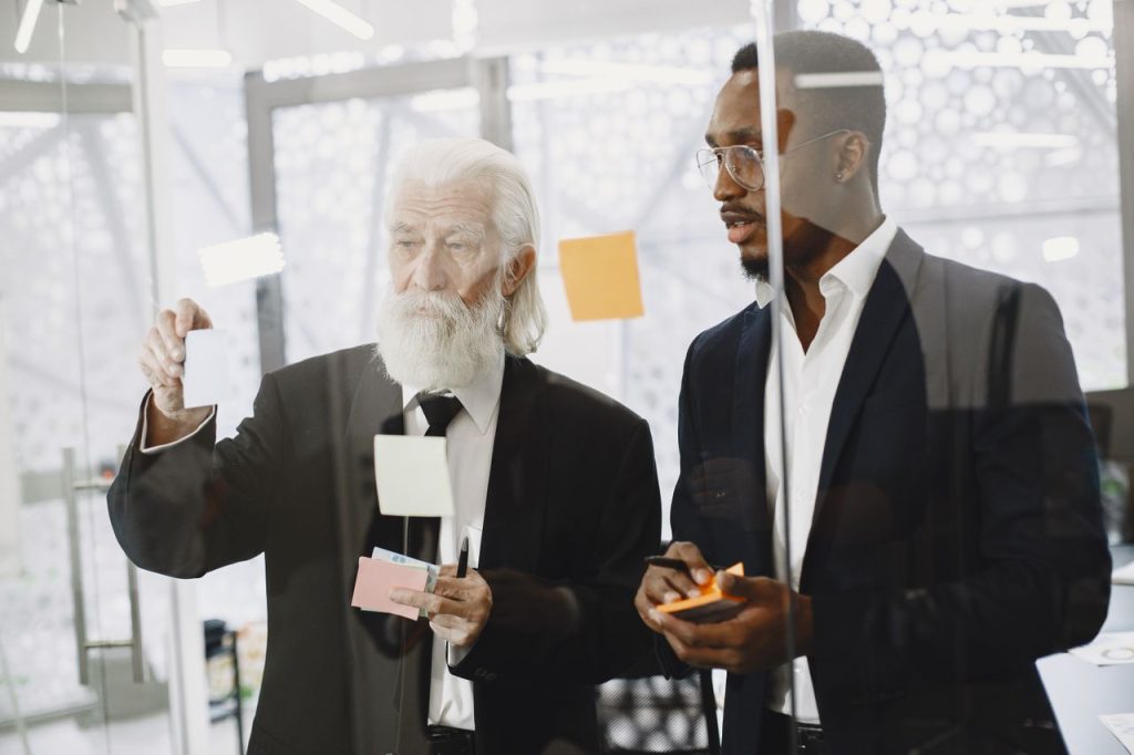 Two men in suits discussing ideas with sticky notes on a glass wall in an office.